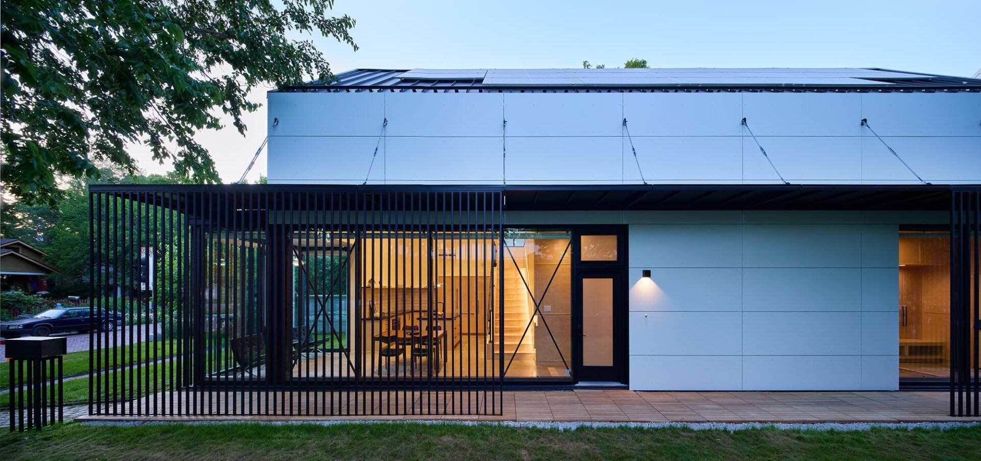 Exterior view of a modern, single-story building at dusk, featuring large glass windows and a flat roof. The building's facade is illuminated by interior lighting and an outdoor wall-mounted lamp.