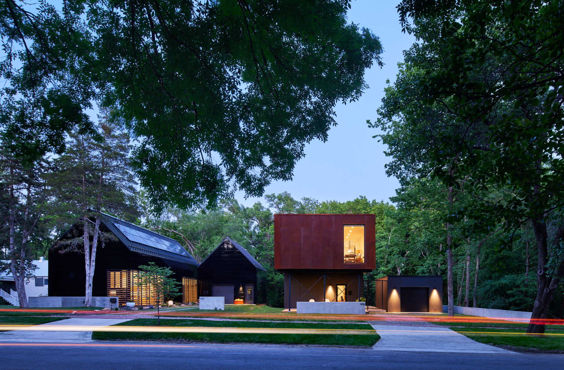 Two modern houses at dusk on a leafy street. 